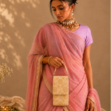 A woman models a light pink saree with a lavender purple short-sleeve blouse, accessorized with gold jewelry and holding a small beige patterned clutch bag. The background is a warm beige wall with leaf shadows, and a vase of baby's breath with a small brass lamp sits on the floor to the left.